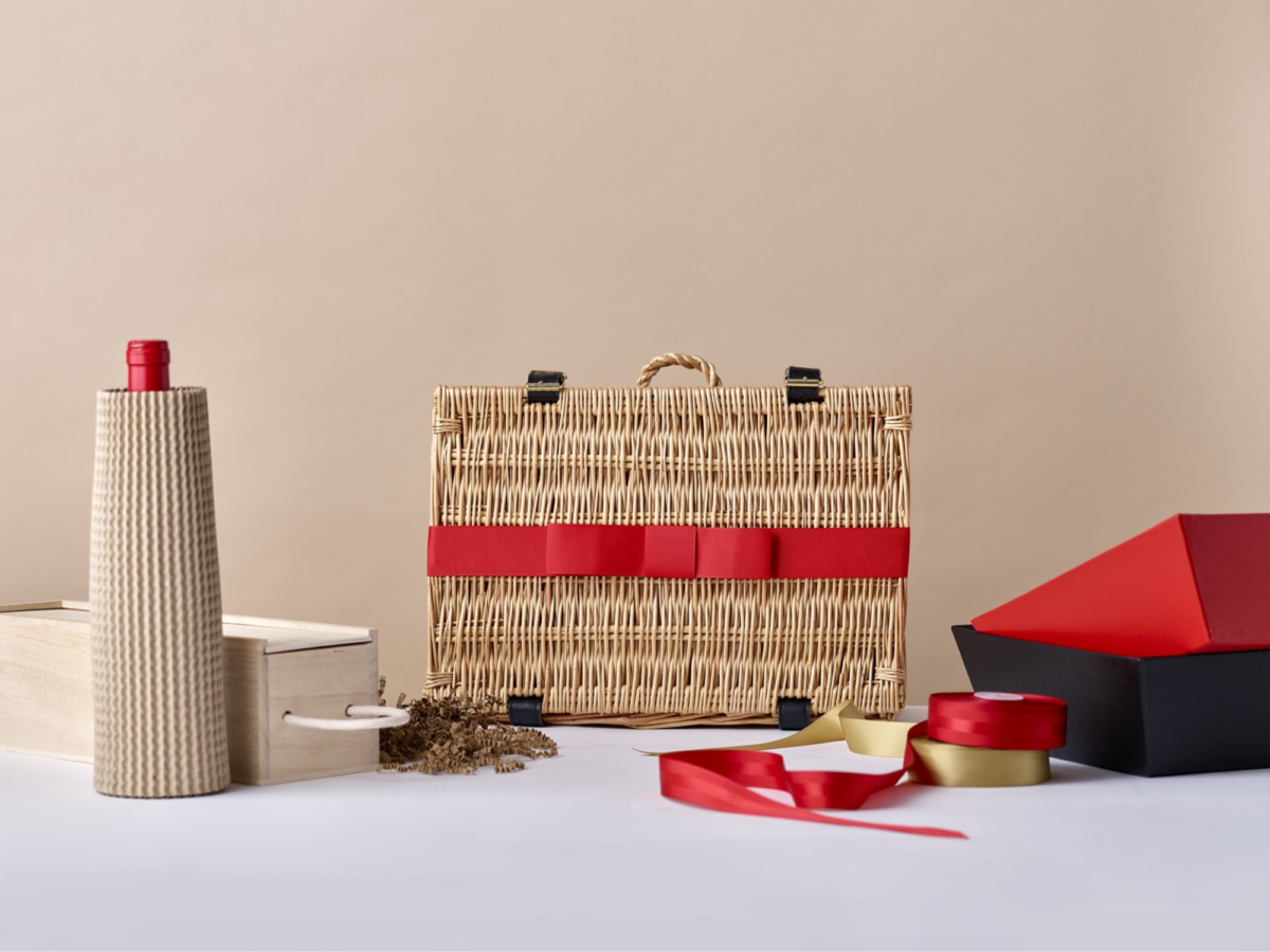 Wicker hamper basket with a red ribbon around it surrounded by hamper making items including ribbon and corrugated card