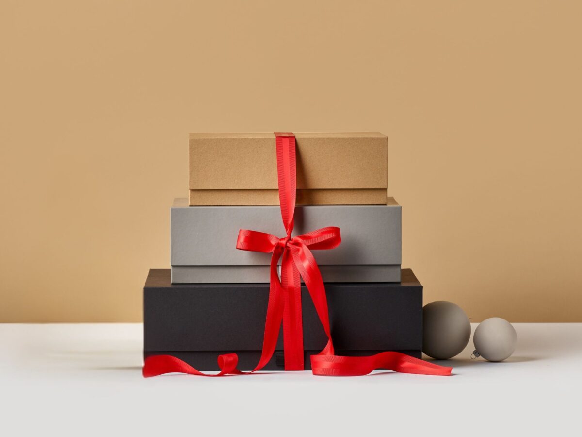 Stack of black, grey and kraft cardboard gift boxes with a red ribbon wrapped around them all and Christmas baubles to the side.