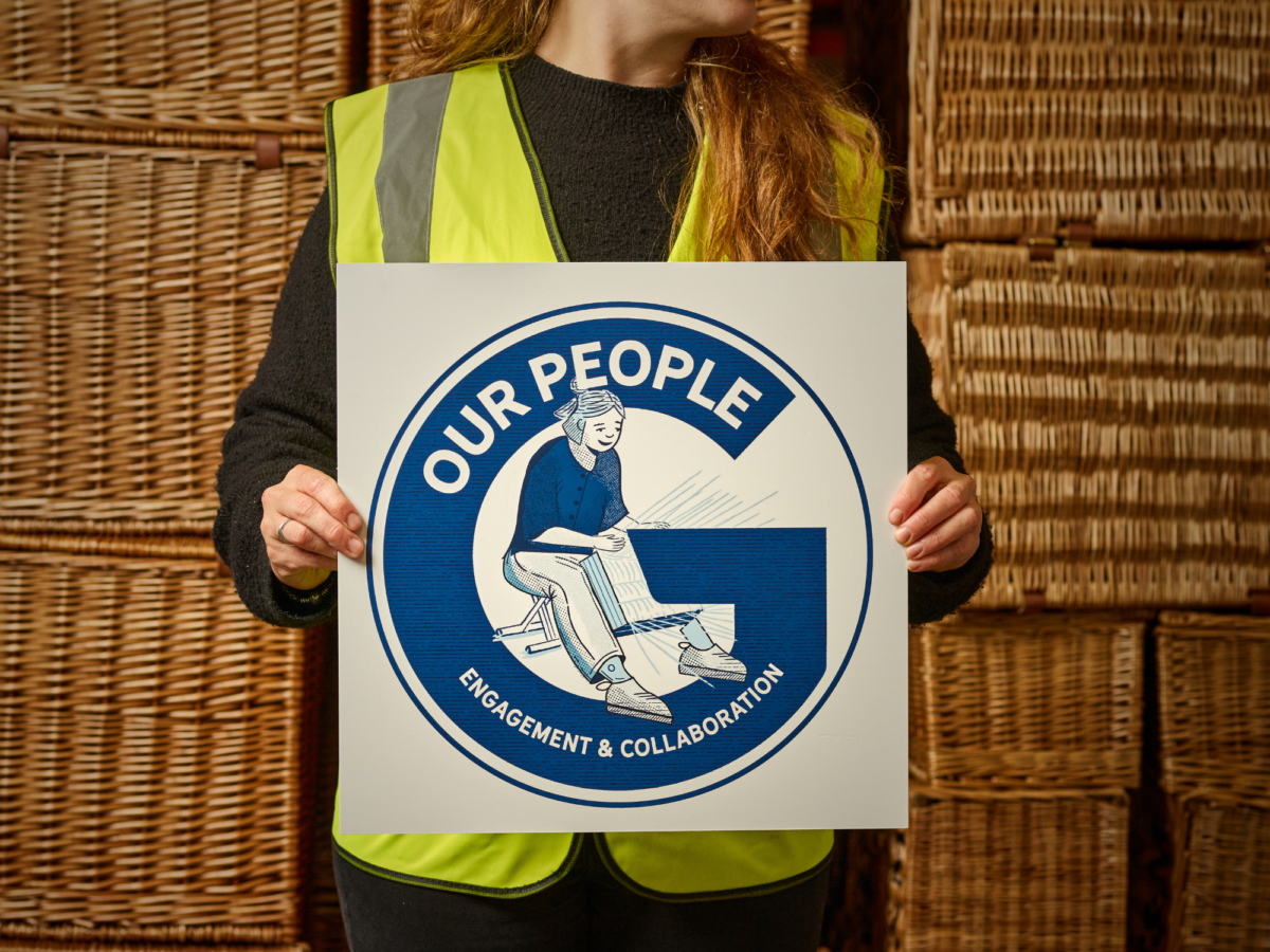 An image of a woman working in a warehouse in front of a stack of wicker hampers holding a sign that says Our People